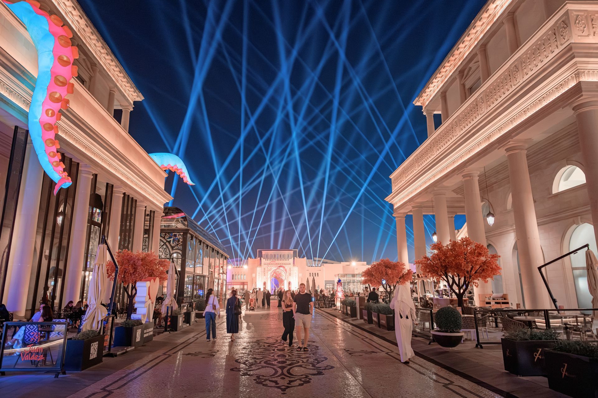 Nighttime pedestrian street with blue laser lights, illuminated buildings, and people walking.