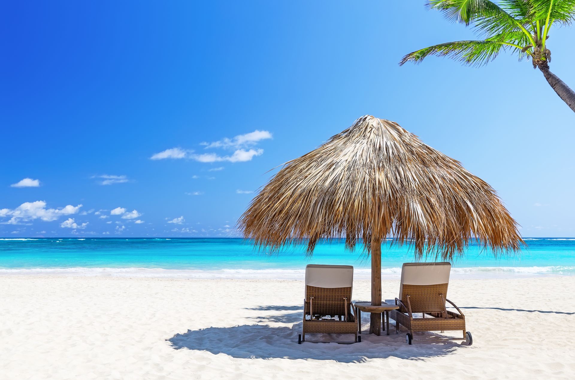 Two lounge chairs under a straw umbrella on a sunny white-sand beach with blue ocean and sky.