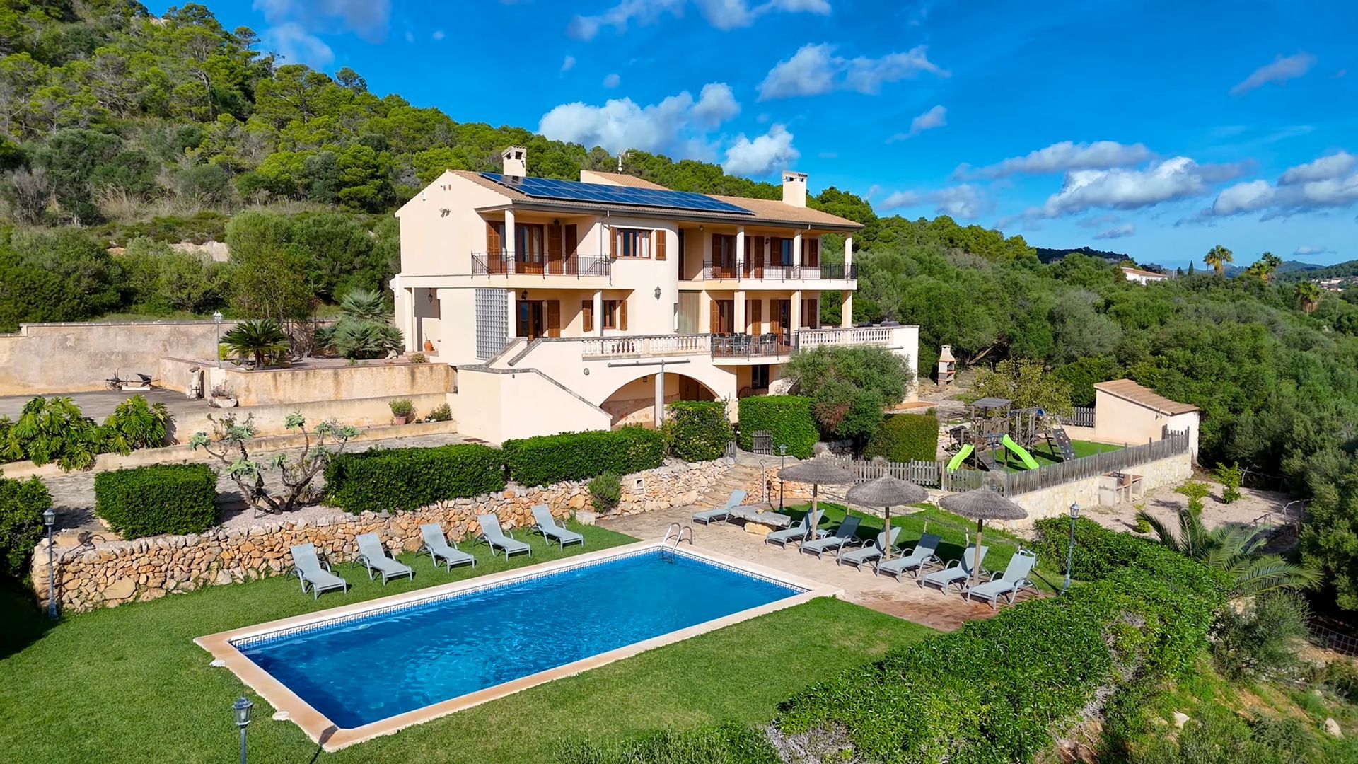 Luxury hillside villa with blue pool, sun loungers, and lush green surroundings under a bright sky
