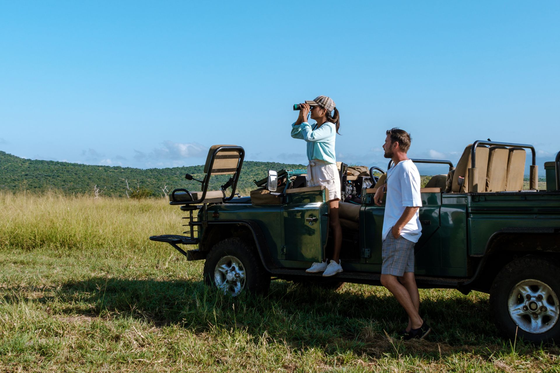 Two people standing by a safari jeep in a grassy field, one looking through binoculars under a blue sky