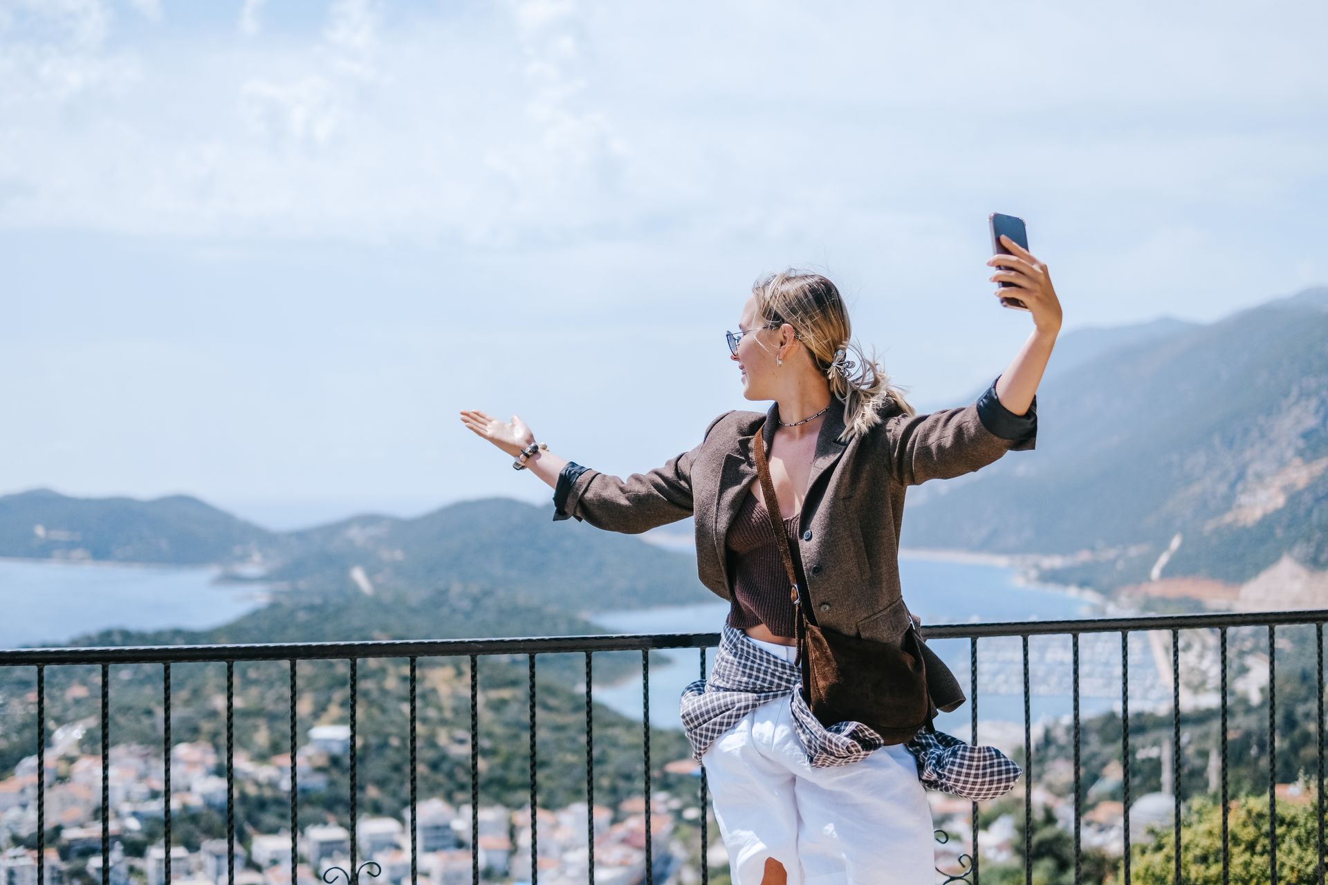 Woman taking a selfie on a balcony with mountains and a coastal town in the background