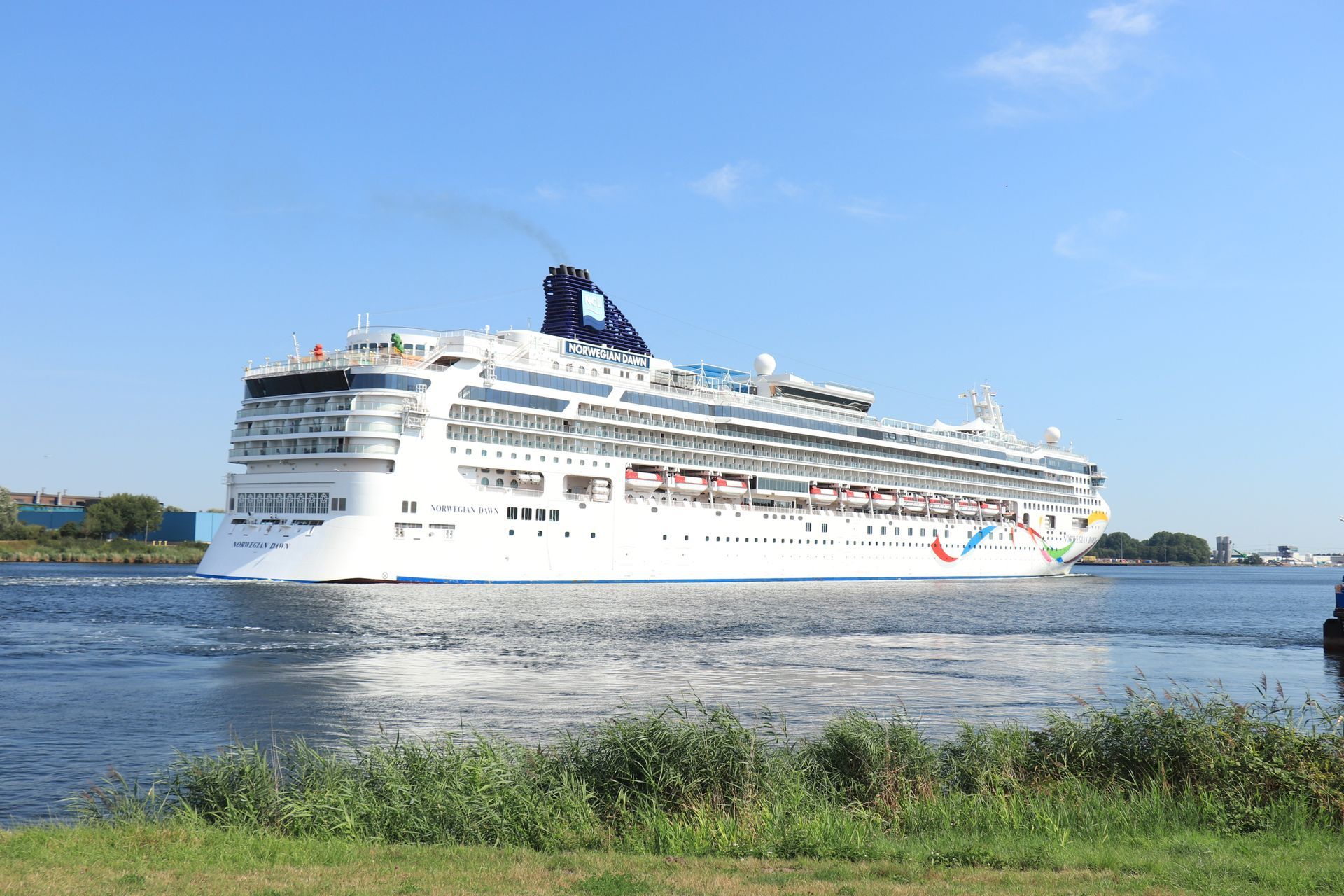 Large white cruise ship on a river under a clear blue sky, viewed from the grassy shoreline
