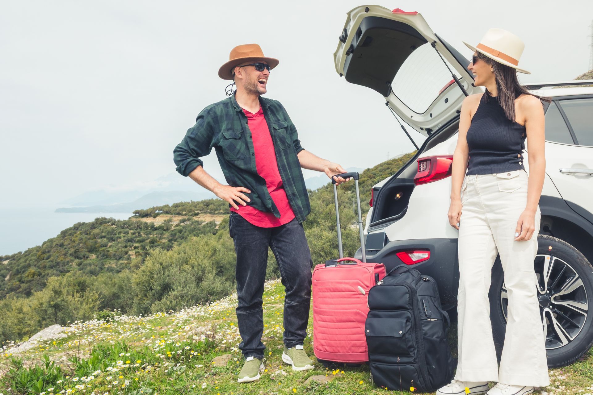 Two hikers with luggage beside an open car trunk on a grassy hillside