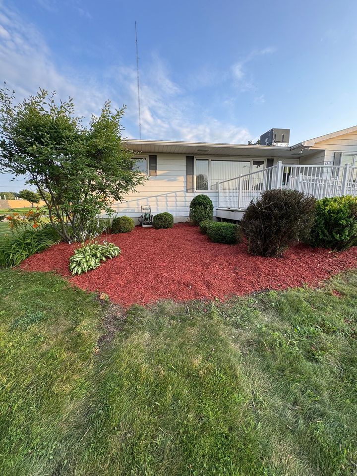 Front yard with white house, red mulch landscaping, green lawn, and a tree under a blue sky
