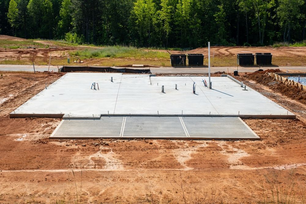 Concrete foundation slab at a construction site, with dirt around it and trees in the background