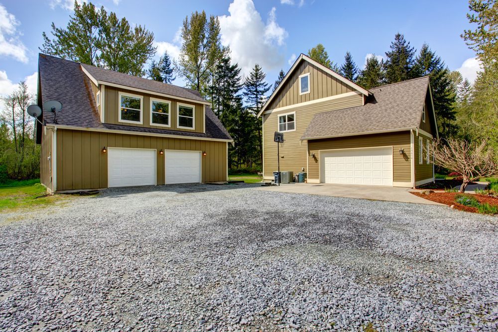 Two tan houses with garages on a gravel driveway, surrounded by trees under a blue sky.