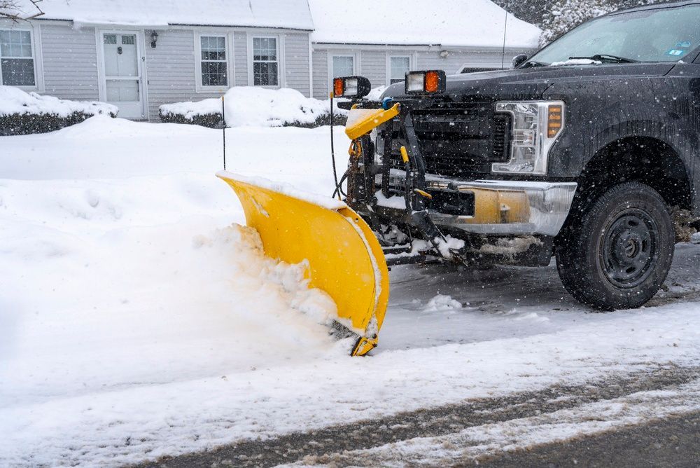 Yellow snowplow clearing a snowy residential street beside parked truck in winter.