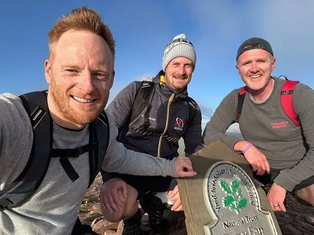 Three hikers smiling on a mountain summit beside a trail marker under a clear sky