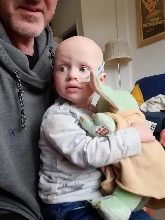 Adult holding a smiling baby with a green plush toy on a couch in a living room