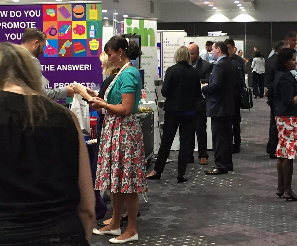 People chatting at an indoor event booth with colorful posters and a woman holding papers in the foreground