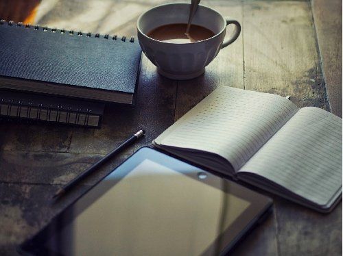 Notebook, tablet, pen, and open book on a desk beside a cup of coffee.