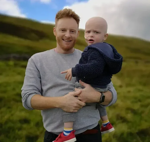 Man holding a child outdoors on a grassy hillside, both facing the camera.