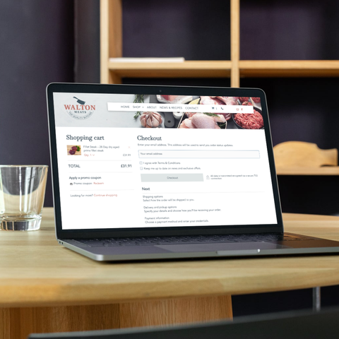 Laptop on a wooden desk displaying a website with product and profile tiles in a bright office setting
