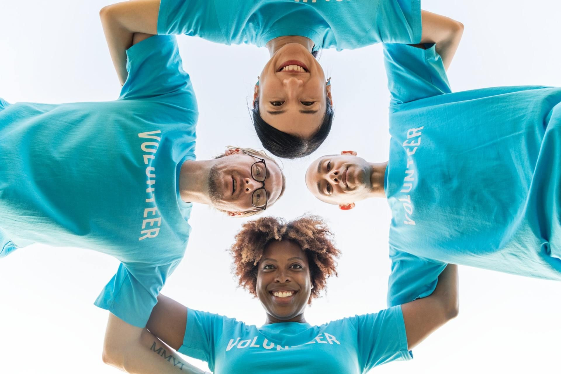 Four smiling people in turquoise shirts lean in a circle, viewed from below.
