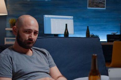 A bald man with a beard is sitting on a couch with a bottle of beer.