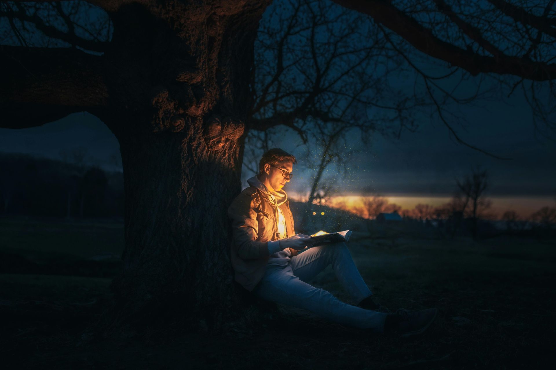 A man is sitting under a tree at night reading a book.