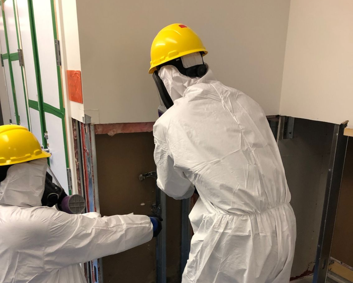 Un homme en combinaison de protection pulvérise une pièce avec un pulvérisateur.