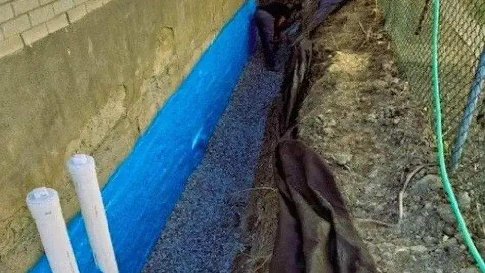 Une membrane étanche bleue longe un mur de terre dans une tranchée étroite, avec une bâche en plastique noir et des tuyaux blancs.