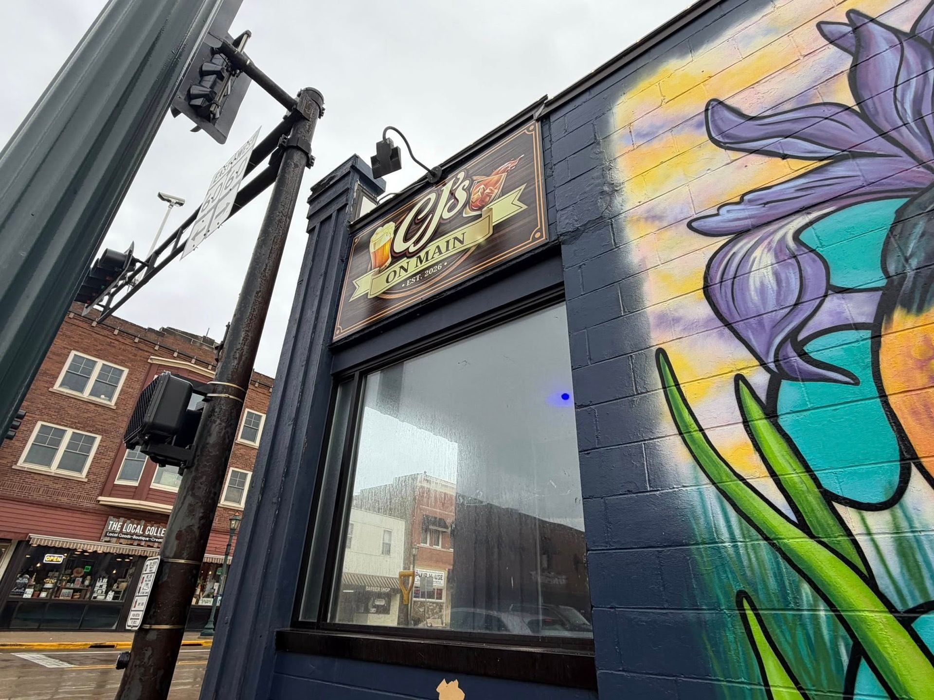 Exterior of CJ's sports bar with a colorful floral mural on a blue brick wall, set in a downtown street intersection.