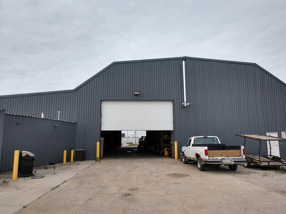Gray industrial warehouse with open garage door, pickup truck, and trash bins in front.
