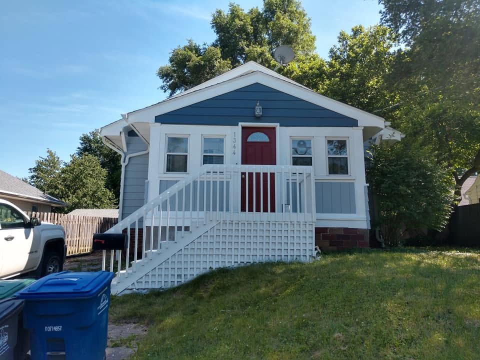 Small blue house with white porch, red front door, and stairs on a grassy hill under a sunny sky