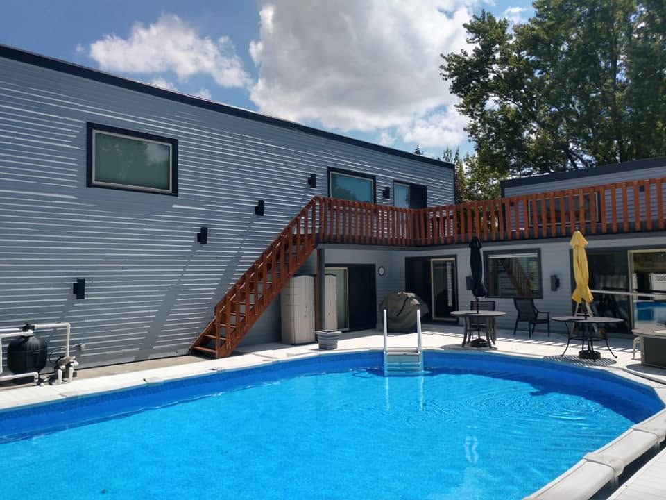 Backyard pool beside a gray two-story house with wooden stairs and patio furniture