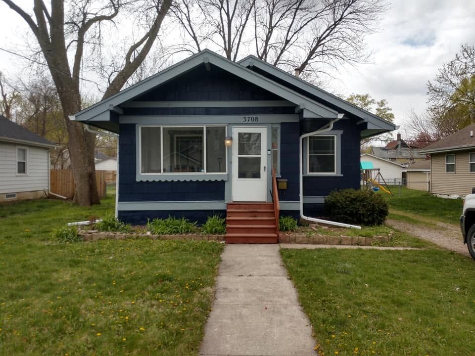 Small dark-blue bungalow with a front porch, red steps, and a central walkway in a residential yard.