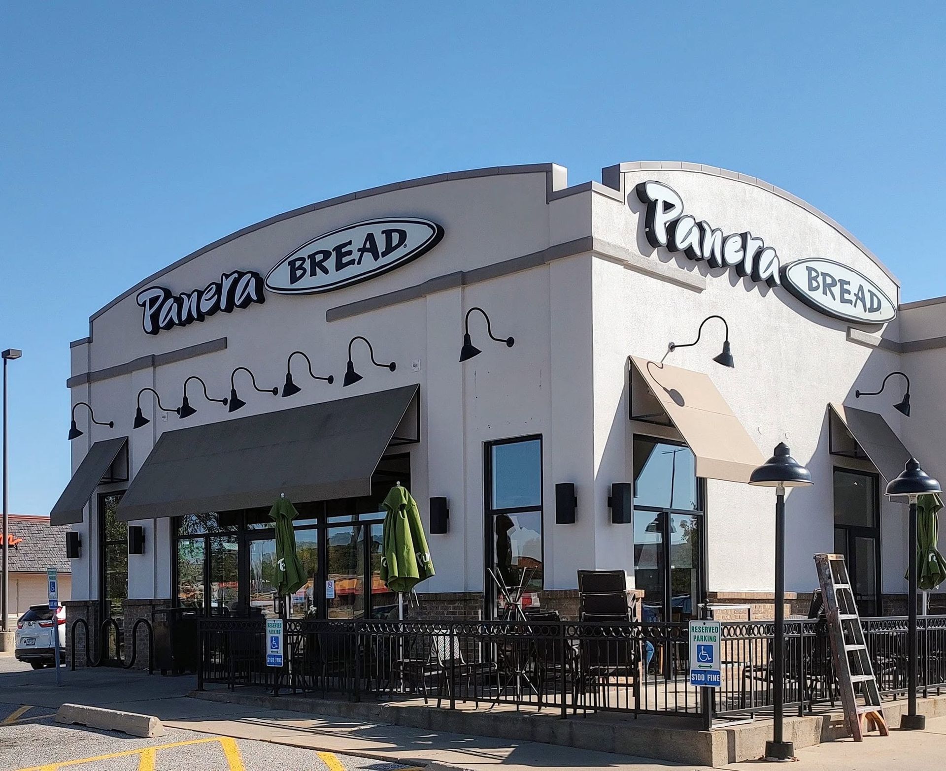 Corner bakery storefront with white stucco walls, black awnings, and outdoor tables under a blue sky.