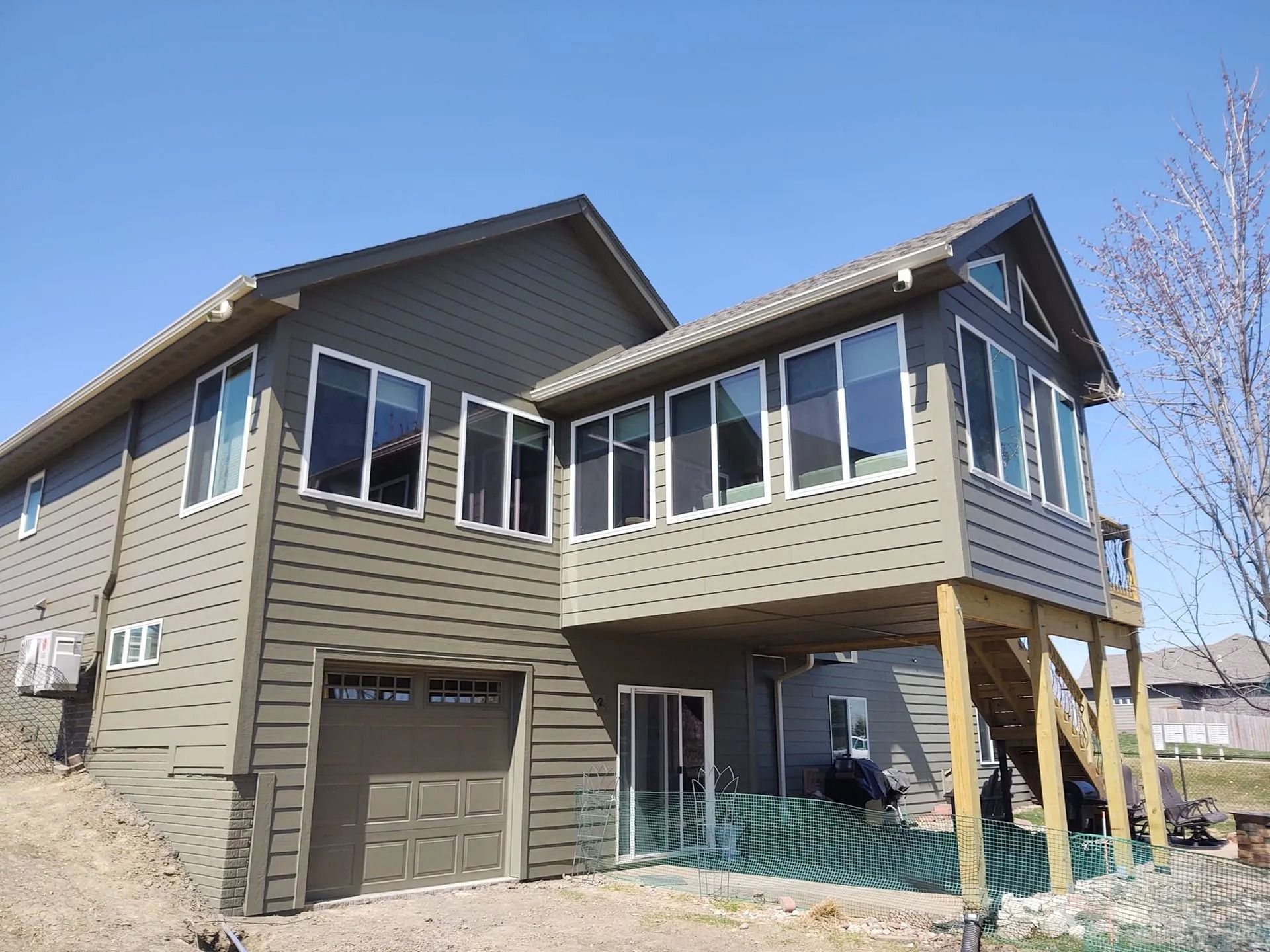 Two-story tan house with a garage, raised deck, and green patio furniture on a sloped lot
