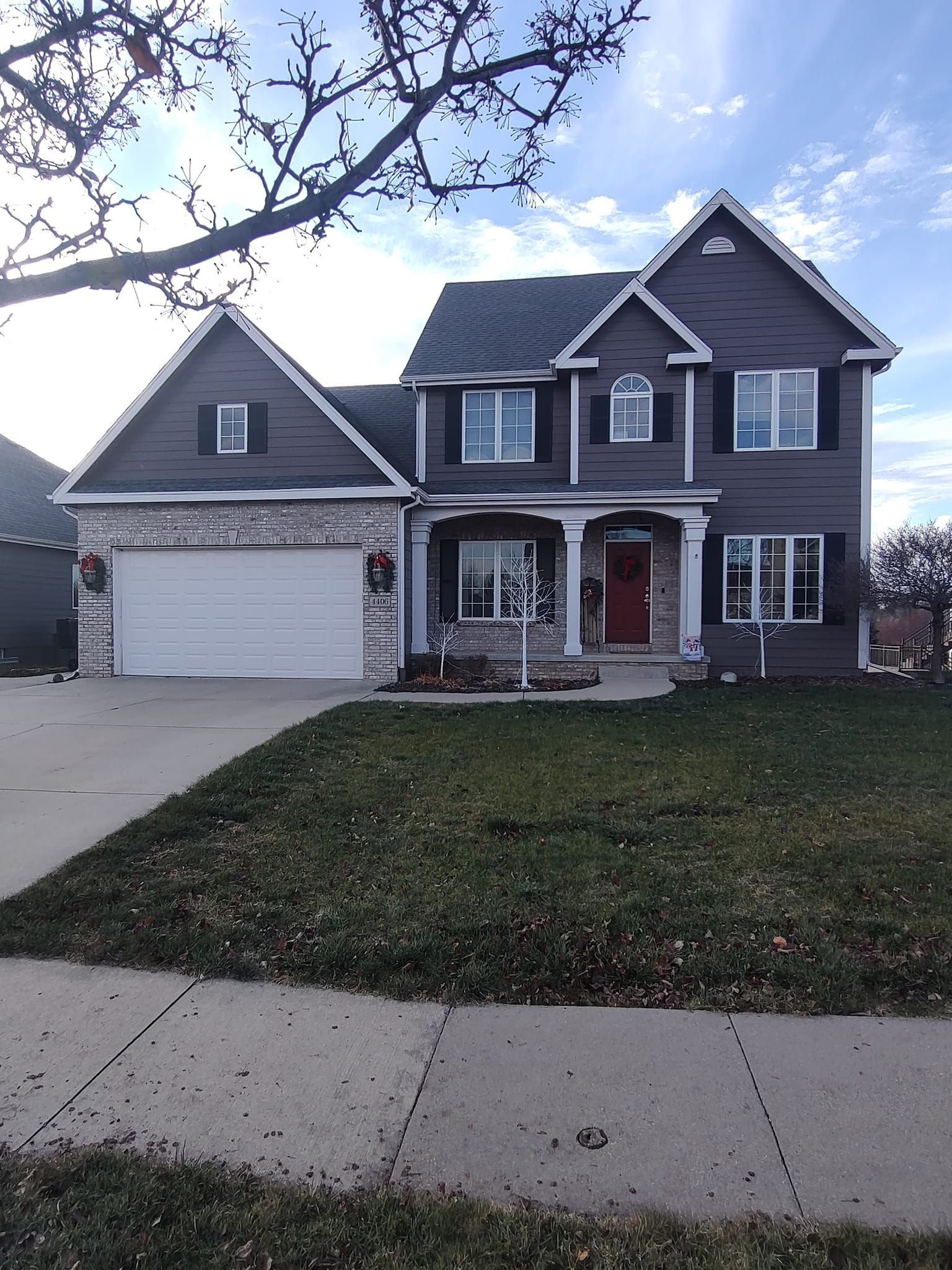 Two-story gray house with white trim, front porch, garage, and a sloped lawn beside a sidewalk.