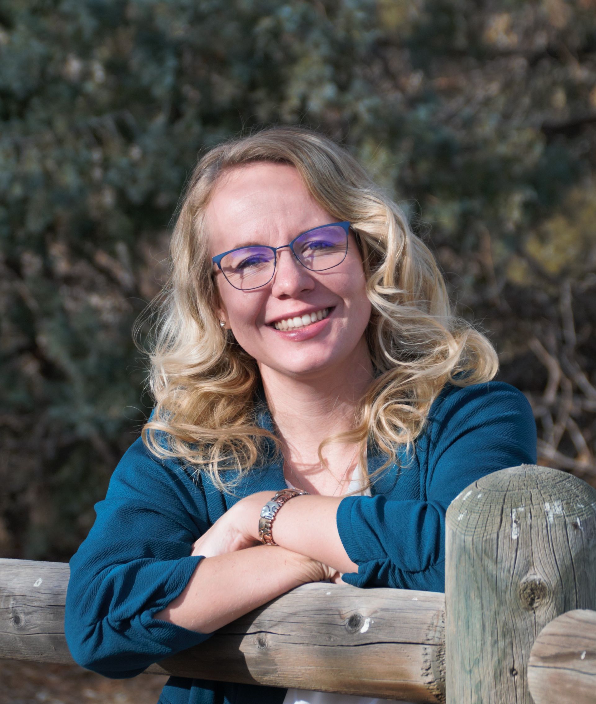 A woman wearing glasses is leaning on a wooden fence and smiling.