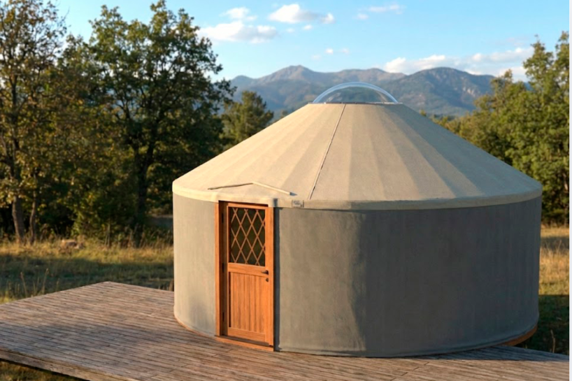 Yurta redonda de color beige con puerta de madera en una terraza, con árboles y montañas de fondo bajo un cielo azul.
