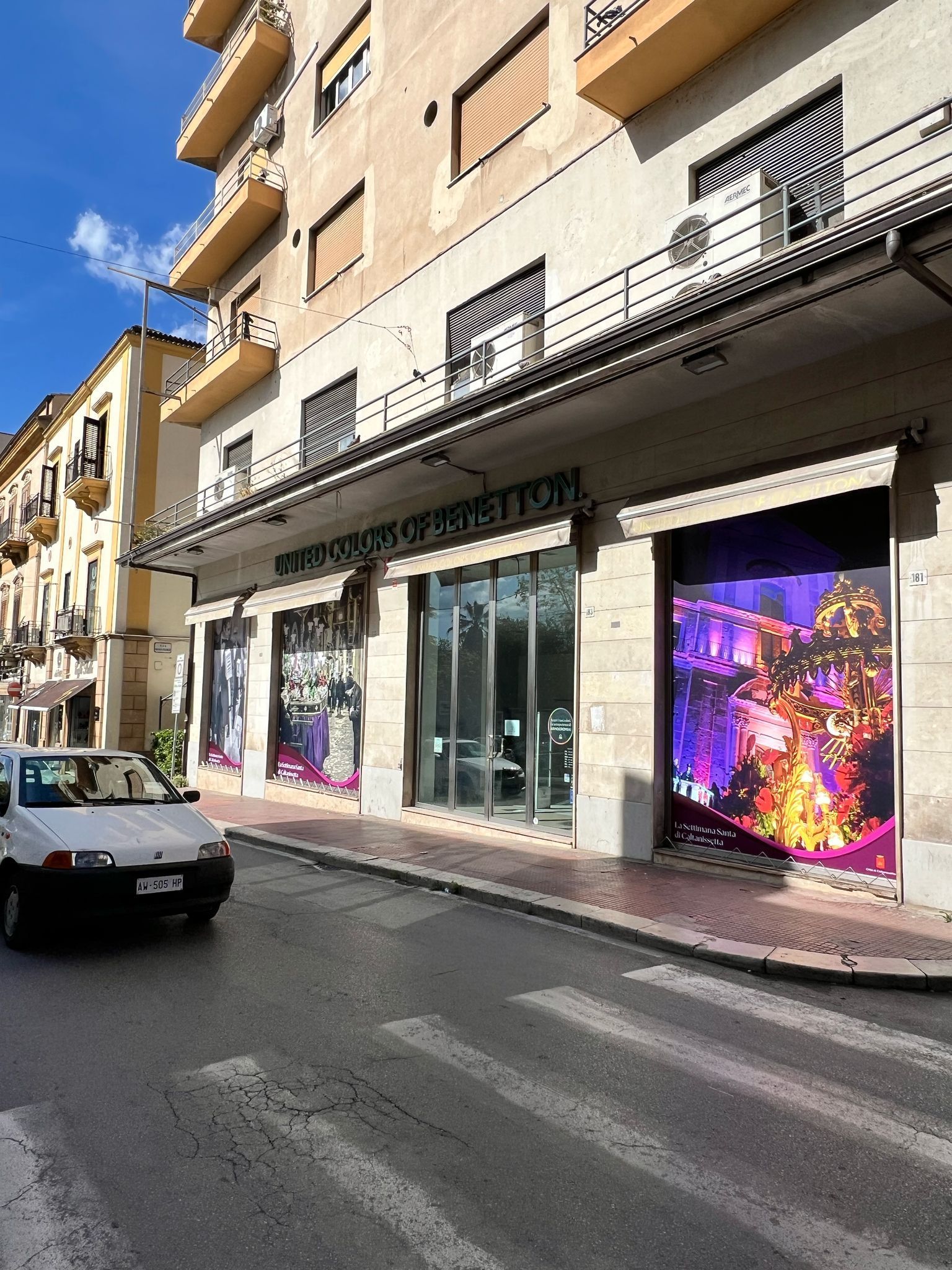 Un'auto bianca passa davanti a un edificio a più piani con vetrine decorate con vivaci luci viola e arancioni.
