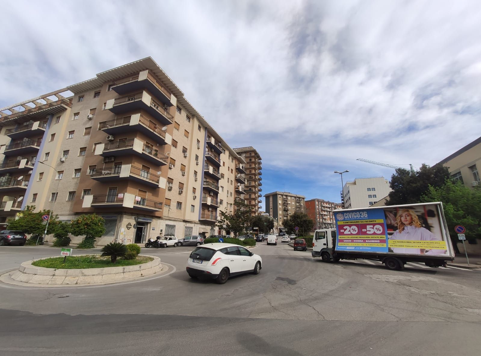 Un'auto bianca e un camion commerciale con una pubblicità attraversano una rotonda vicino a dei palazzi residenziali.