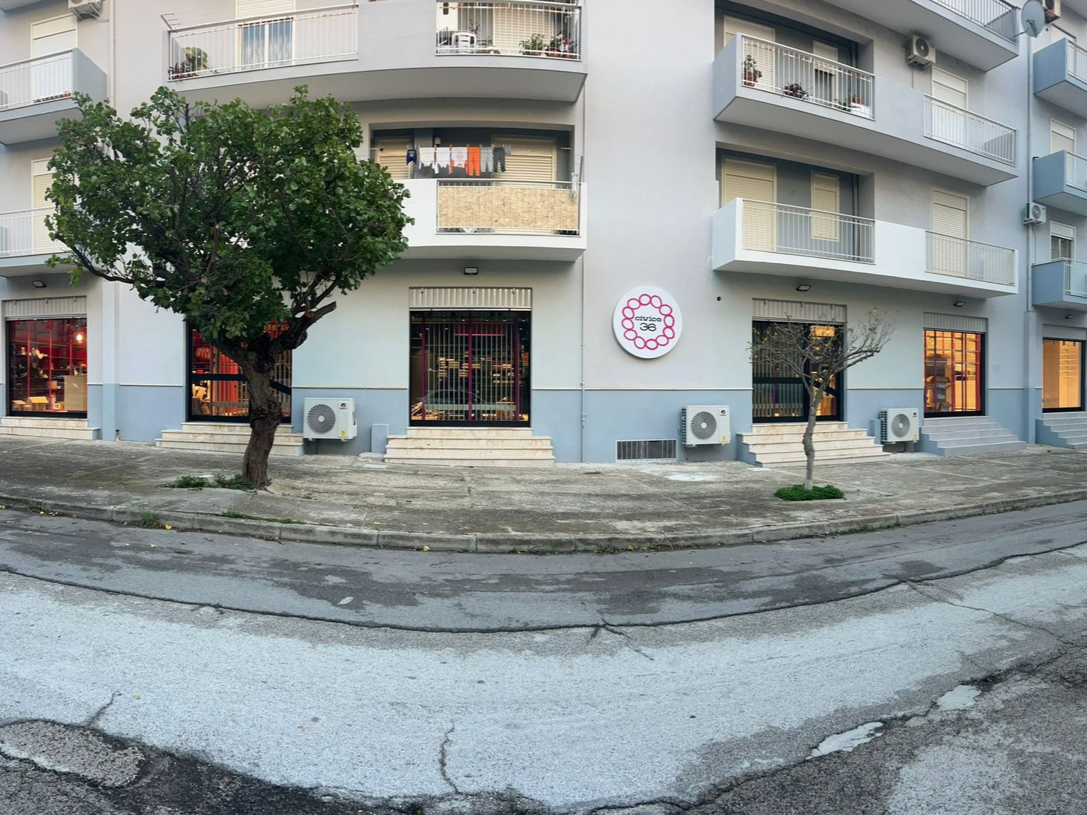 Un edificio residenziale a più piani con negozi al piano terra, un albero di fronte e un'insegna circolare bianca sul muro.