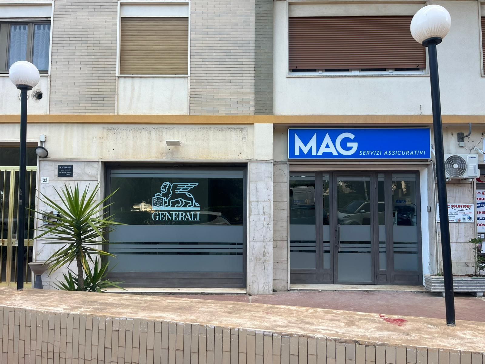Le vetrine di un'agenzia assicurativa Generali e di un ufficio immobiliare MAG su una soleggiata via cittadina.