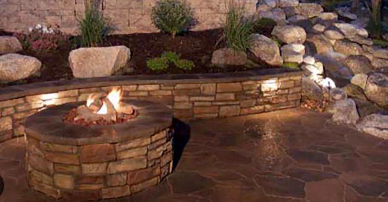 Stone fire pit with flames in a landscaped outdoor patio at dusk