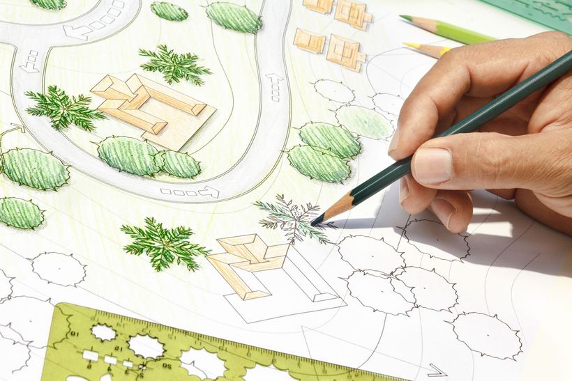 Hand sketching a landscape plan with green trees, paths, and buildings on paper