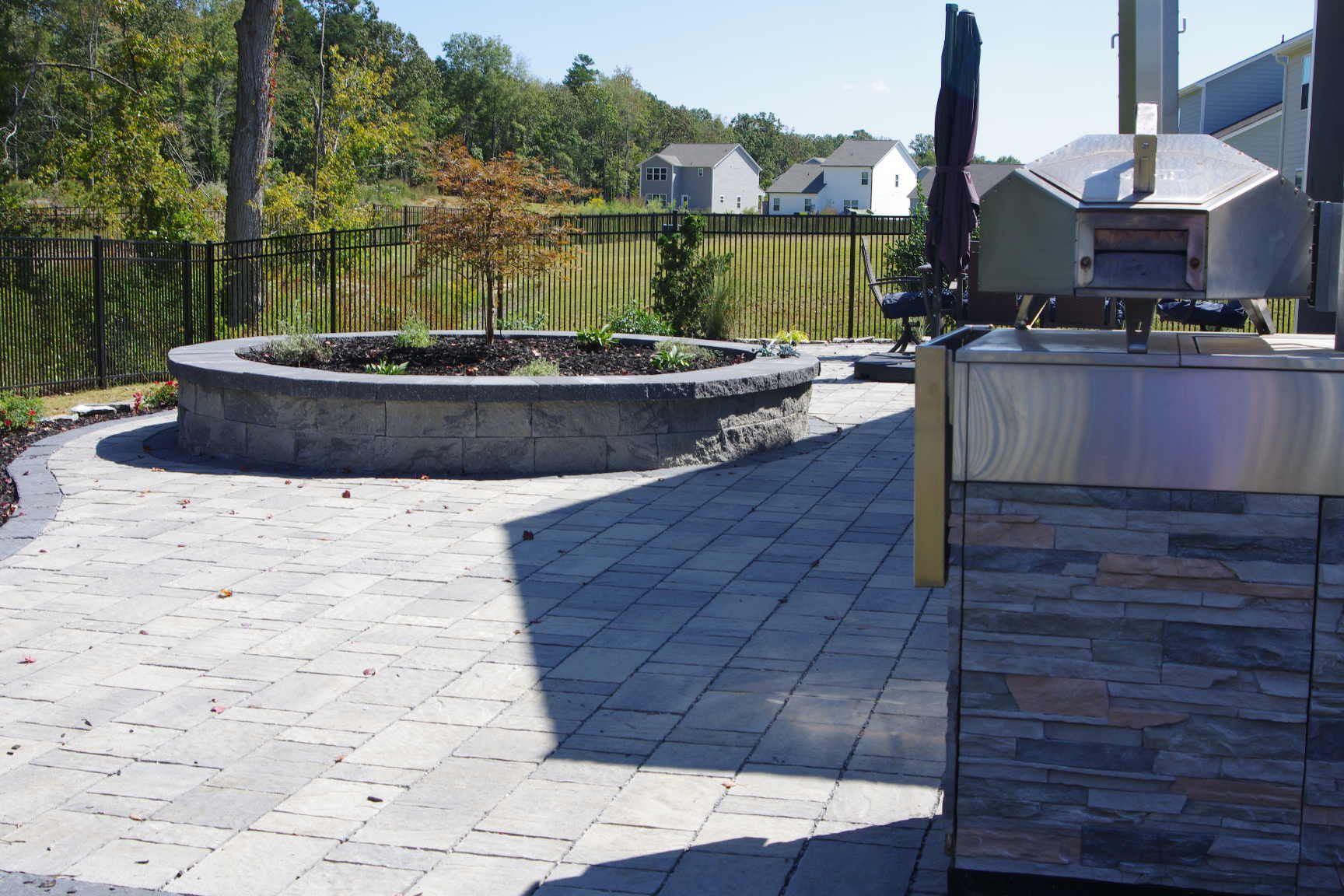 Stone patio with a curved raised planter and an outdoor grill in a sunny backyard.