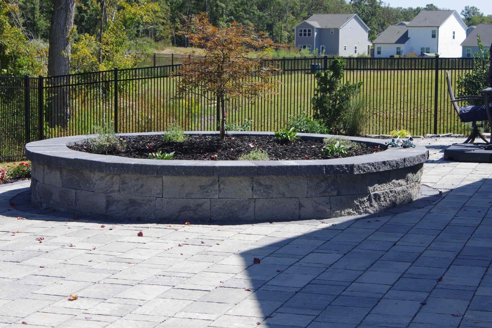 Circular stone planter in a paved patio, with a fence and houses in the background