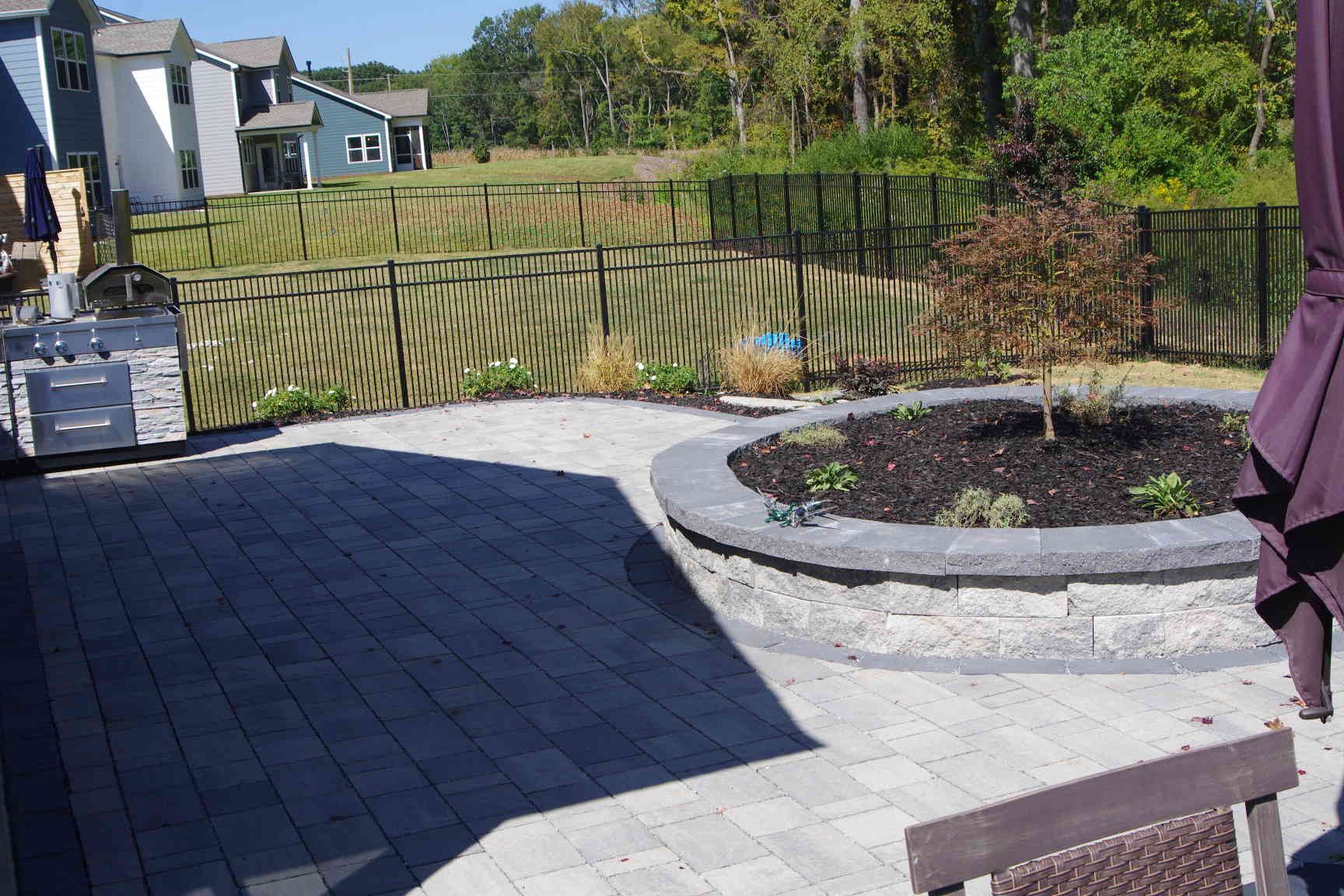 Backyard patio with stone pavers, raised garden bed, and wooden fence on a sunny day