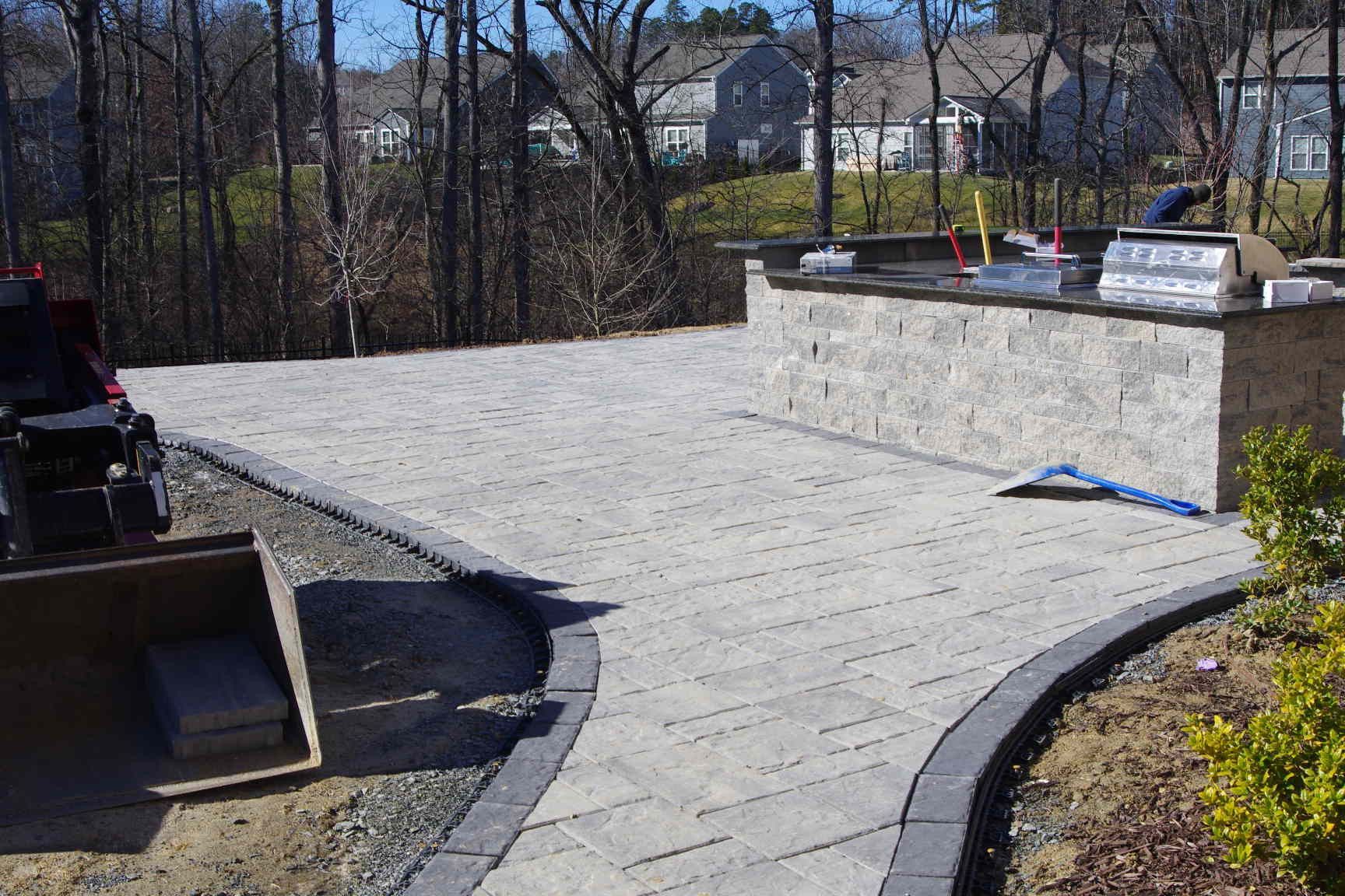Stone patio with curved walkway, raised wall, and outdoor kitchen in a sunny backyard
