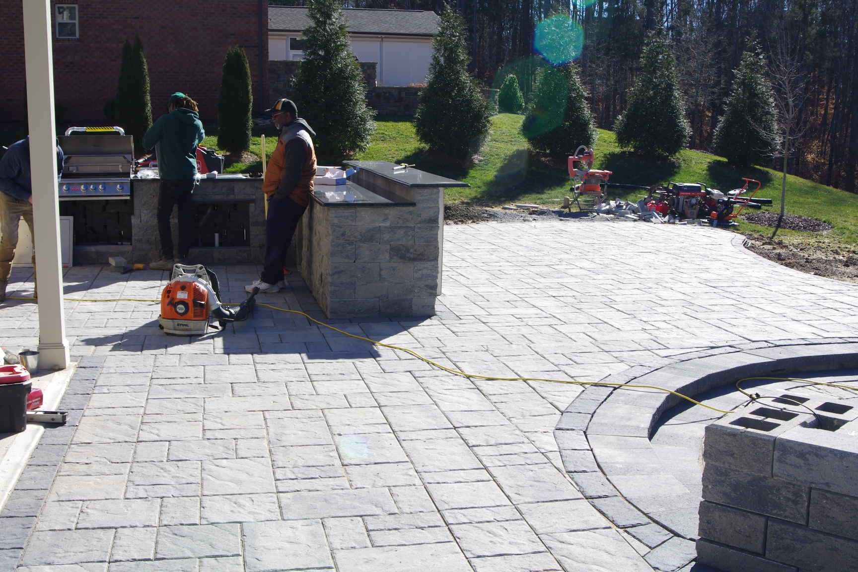 Two workers installing stone pavers in a sunlit courtyard with a leaf blower nearby