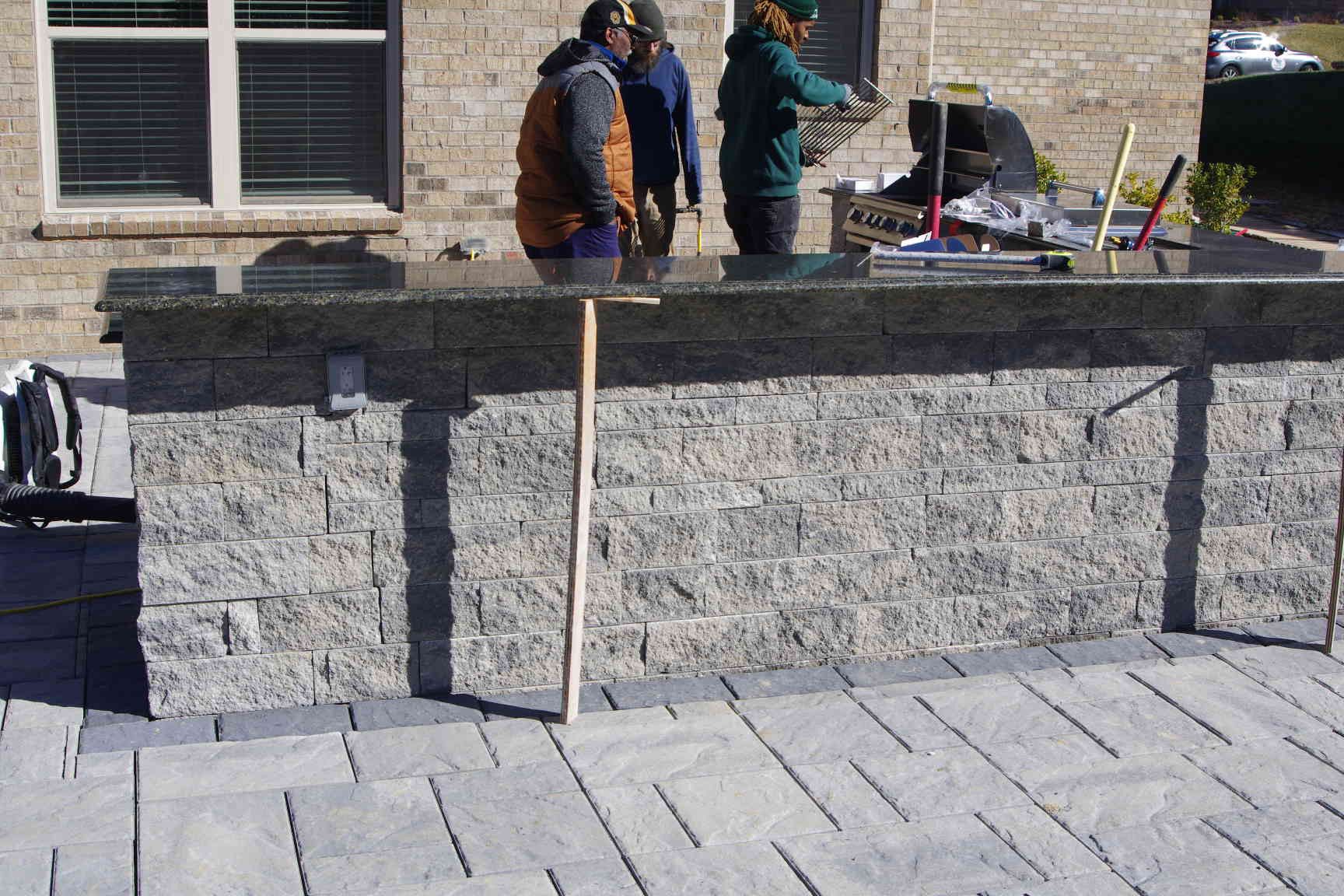 Workers building a stone wall beside a sidewalk, with tools and materials nearby.