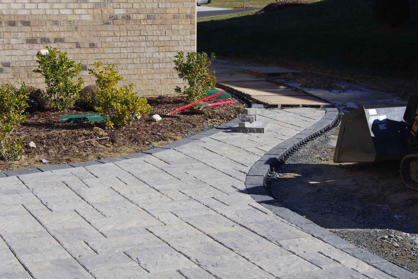 Curved light-gray paver walkway beside a brick wall and landscaped bed, ending at a driveway.