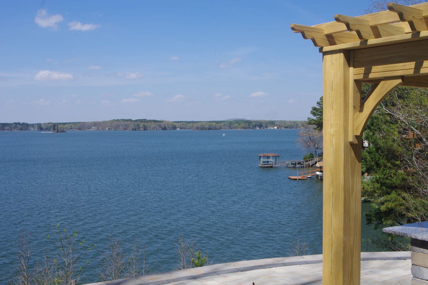 Blue lake view from a wooden deck with a pergola, shoreline in the distance, and a small dock.