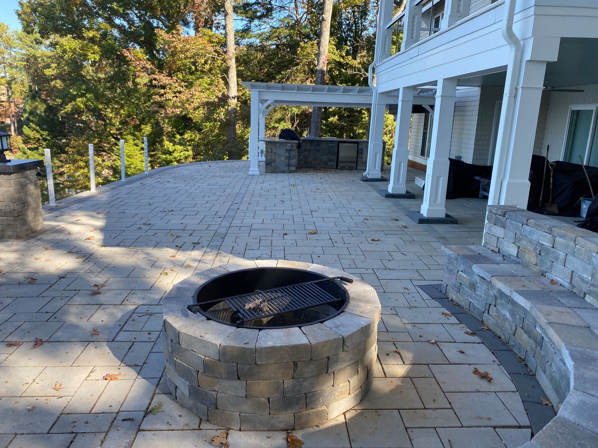 Stone patio with a circular fire pit beside a white porch and steps, backed by trees