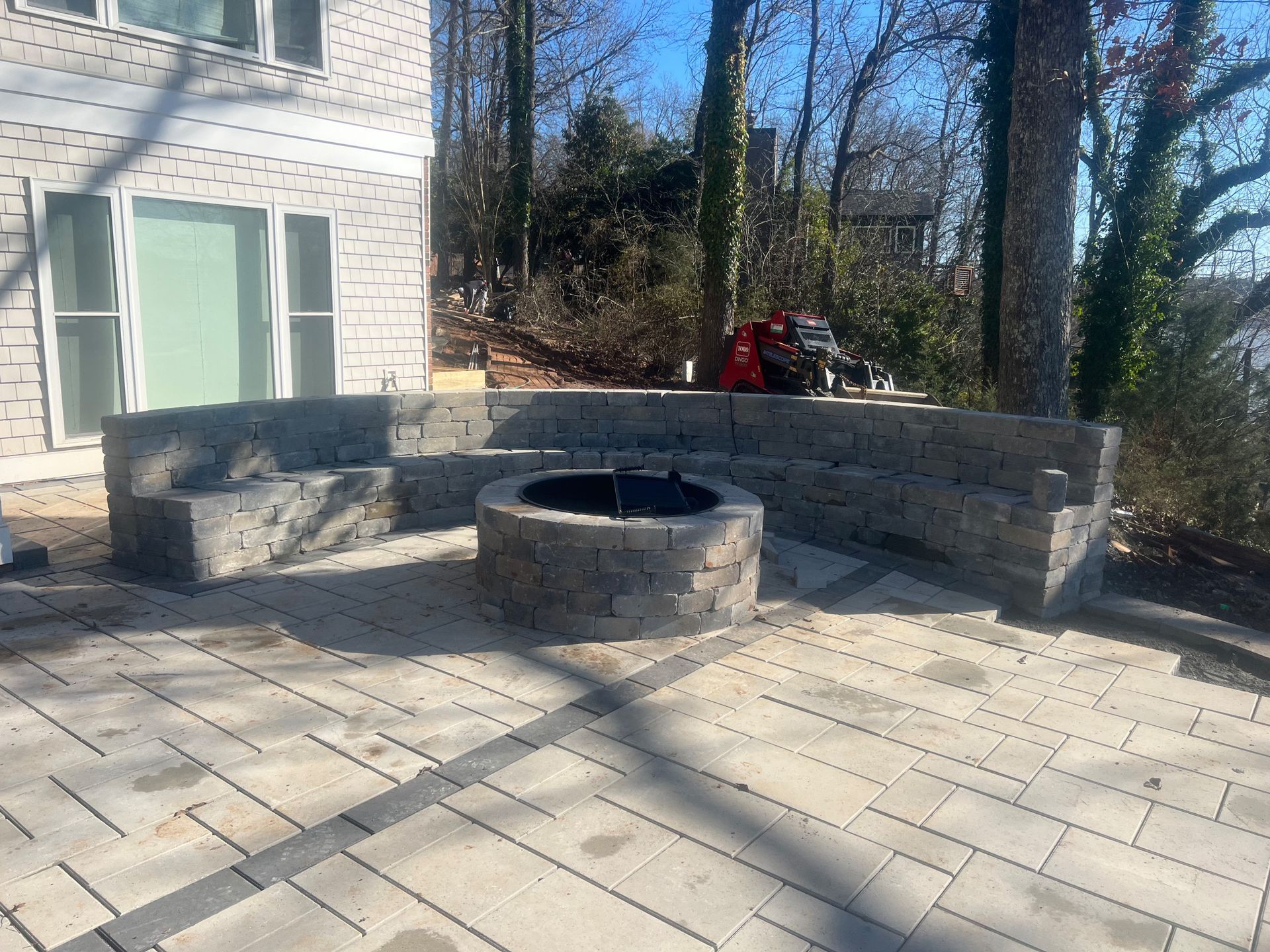 Stone patio with circular fire pit, built-in seating, and a house in the background on a sunny day