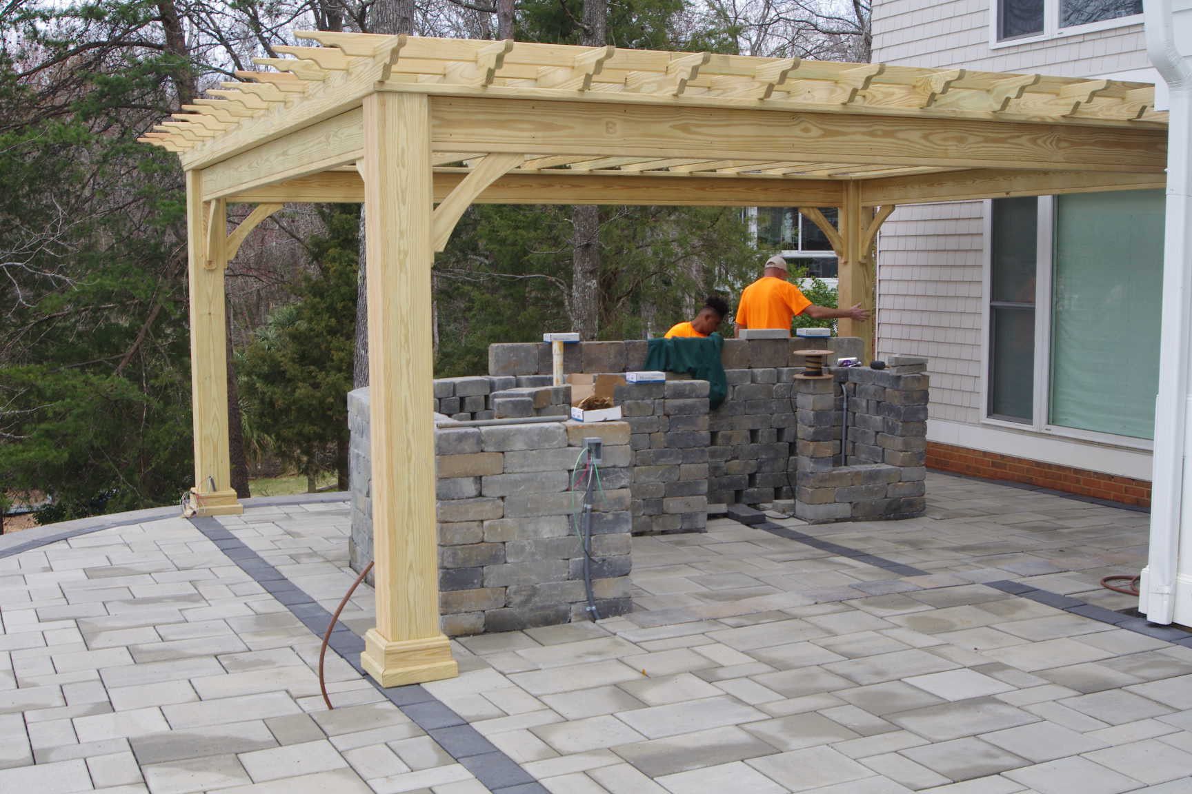 Two workers in orange shirts building an outdoor stone fireplace under a wooden pergola on a paved patio