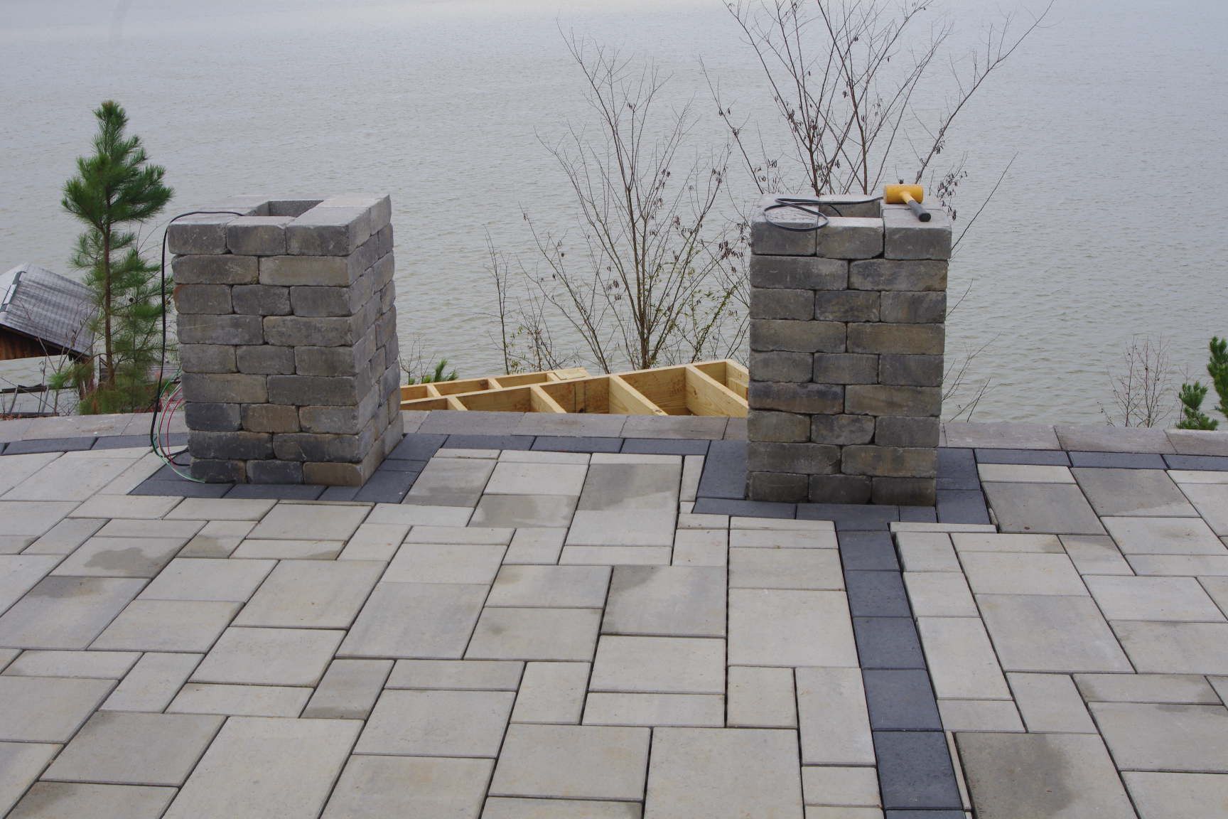 Stone patio with two short brick pillars beside a calm waterfront and leafless shrubs
